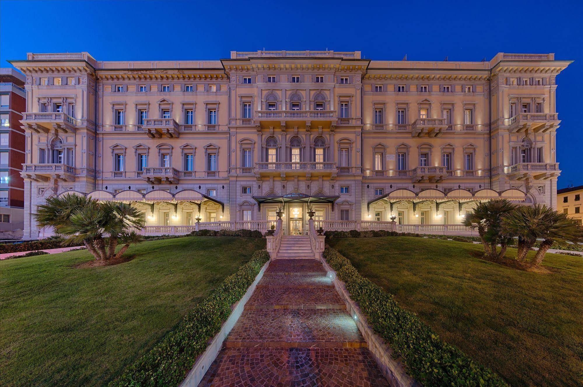 Gh Palazzo Suite & Spa Livorno Ngoại thất bức ảnh The photo shows a large, elegant building, likely a hotel or historic structure, illuminated at dusk. It features a classic architectural style with multiple balconies and ornate detailing on the façade. The building is framed by well-maintained gree